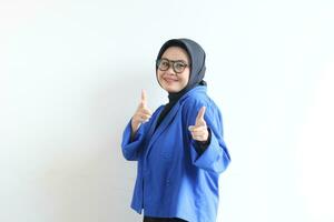 Beautiful young Asian Muslim woman in glasses, hijab and wearing blue blazer smiling confident and excited with both hands pointing forward photo