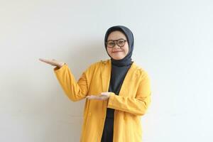 Beautiful young Asian Muslim woman in glasses, hijab and wearing yellow blazer smiling confident and happy with both hands presenting empty space photo