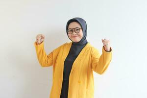 beautiful young Asian Muslim woman, wearing glasses and yellow blazer with strong gesture by lifting her arms and muscles photo