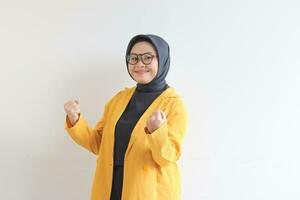 beautiful young Asian Muslim woman, wearing glasses and yellow blazer with happy expression while clenching fist photo