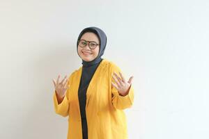 Young beautiful Asian Muslim woman, wearing glasses and yellow blazer with happy smiling face expression photo