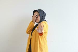 Beautiful young Asian Muslim woman, wearing glasses and yellow blazer covering 1 side of face with hand photo