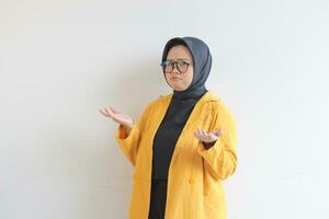 Beautiful young Asian Muslim woman in glasses, hijab and wearing yellow blazer with confused expression and both of hands presenting empty space photo