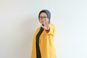 Beautiful young Asian Muslim woman, wearing glasses and yellow blazer showing stop gesture photo