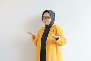 Beautiful young Asian Muslim woman in glasses, hijab and wearing yellow blazer with confused expression and both of hands presenting empty space photo