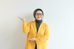 Beautiful young Asian Muslim woman, wearing glasses and yellow blazer showing palms while smiling photo