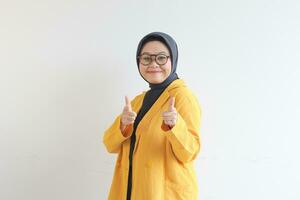 Beautiful young Asian Muslim woman in glasses, hijab and wearing yellow blazer smiling confident and excited with both hands pointing forward photo