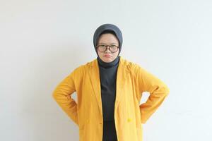 Young beautiful Asian Muslim woman, wearing glasses and yellow blazer with angry facial expression photo