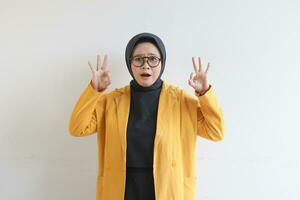 beautiful young Asian Muslim woman, wearing glasses and yellow blazer with hand gesture of approval or OK photo