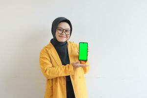 beautiful young asian woman in glasses, hijab and wearing yellow blazer showing green screen mobile phone while smiling photo