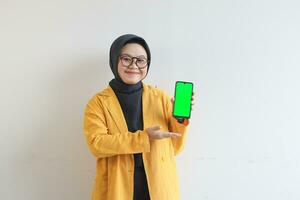 beautiful young asian woman in glasses, hijab and wearing yellow blazer showing green screen mobile phone while smiling photo