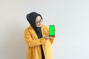 beautiful young asian woman in glasses, hijab and wearing yellow blazer showing green screen mobile phone while smiling photo