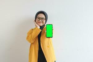 beautiful young asian woman in glasses, hijab and wearing yellow blazer showing green screen mobile phone while smiling photo