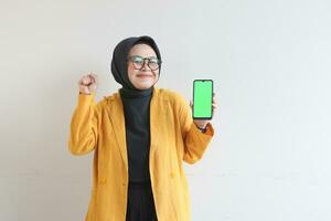 beautiful young asian woman in glasses, hijab and wearing yellow blazer is holding green screen mobile phone with happy face photo