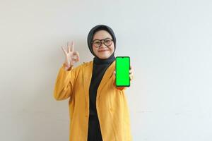 beautiful young asian woman in glasses, hijab and wearing yellow blazer showing green screen mobile phone while smiling photo