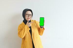 beautiful young asian woman in glasses, hijab and wearing yellow blazer is holding green screen mobile phone with sullen face photo