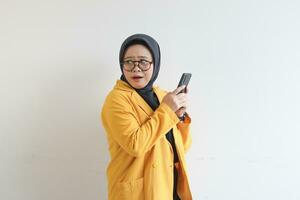 hermosa joven asiático musulmán mujer, vistiendo lentes y amarillo chaqueta de sport mirando oblicuo mientras participación móvil teléfono foto