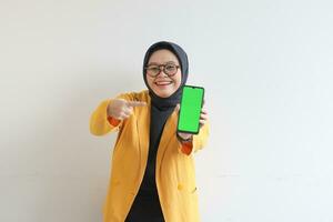 beautiful young asian woman in glasses, hijab and wearing yellow blazer is holding green screen mobile phone with happy face photo