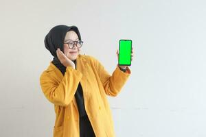 beautiful young asian woman in glasses, hijab and wearing yellow blazer showing green screen mobile phone while smiling photo