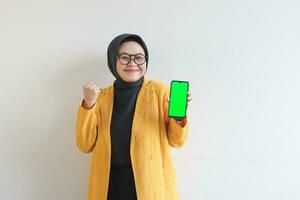 beautiful young asian woman in glasses, hijab and wearing yellow blazer showing green screen mobile phone while smiling photo