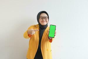 beautiful young asian woman in glasses, hijab and wearing yellow blazer is holding green screen mobile phone with happy face photo