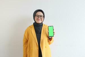 beautiful young asian woman in glasses, hijab and wearing yellow blazer is holding green screen mobile phone with happy face photo