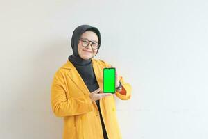 beautiful young asian woman in glasses, hijab and wearing yellow blazer is holding green screen mobile phone with happy face photo