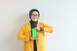 beautiful young asian woman in glasses, hijab and wearing yellow blazer is holding green screen mobile phone with happy face photo