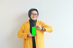 beautiful young asian woman in glasses, hijab and wearing yellow blazer is holding green screen mobile phone with happy face photo