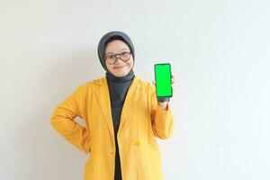 beautiful young asian woman in glasses, hijab and wearing yellow blazer is holding green screen mobile phone with happy face photo