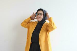 Beautiful young Asian Muslim woman, wearing glasses and yellow blazer showing stress gesture while holding head photo