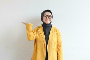 hermosa joven asiático musulmán mujer en anteojos, hijab y vistiendo amarillo chaqueta de sport sonriente confidente y contento con mano presentación vacío espacio foto