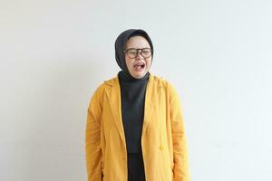 Young beautiful Asian Muslim woman, wearing glasses and yellow blazer with angry facial expression photo