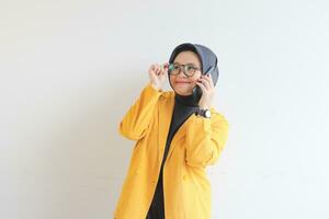 portrait of beautiful asian woman in hijab, glasses and wearing yellow blazer making phone call while smiling photo