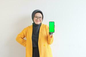 beautiful young asian woman in glasses, hijab and wearing yellow blazer is holding green screen mobile phone with happy face photo