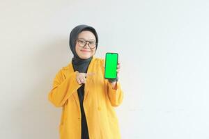 beautiful young asian woman in glasses, hijab and wearing yellow blazer is holding green screen mobile phone with happy face photo