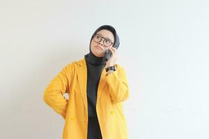 portrait of beautiful asian woman in hijab, glasses and wearing yellow blazer making phone call while thinking photo