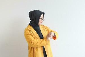 hermosa joven asiático musulmán mujer, vistiendo lentes y amarillo chaqueta de sport gesticulando a reloj foto