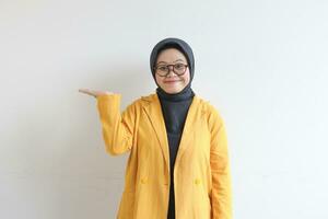 Beautiful young Asian Muslim woman in glasses, hijab and wearing yellow blazer smiling confident and happy with hand presenting empty space photo