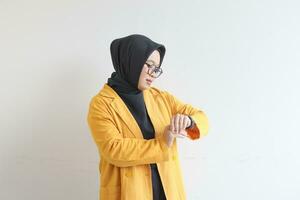 hermosa joven asiático musulmán mujer, vistiendo lentes y amarillo chaqueta de sport gesticulando a reloj foto