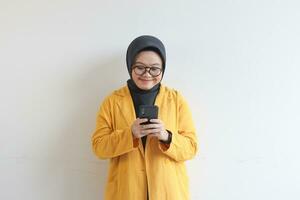 beautiful young asian woman in glasses, hijab and wearing yellow blazer looking at her mobile phone with happy face photo