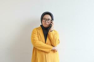 portrait of beautiful asian woman in hijab, glasses and wearing yellow blazer making phone call while looking sideways photo