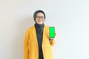 beautiful young asian woman in glasses, hijab and wearing yellow blazer is holding green screen mobile phone with happy face photo