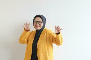 Beautiful young Asian Muslim woman, wearing glasses and yellow blazer making angry gestures while laughing photo