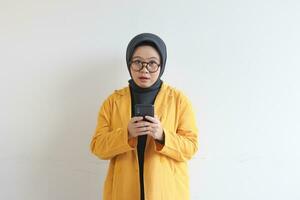 beautiful young asian woman in glasses, hijab and wearing yellow blazer looking at camera while holding mobile phone with shocked face photo