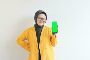 beautiful young asian woman in glasses, hijab and wearing yellow blazer is holding green screen mobile phone with happy face photo