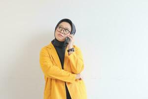 portrait of beautiful asian woman in hijab, glasses and wearing yellow blazer making phone call while thinking photo