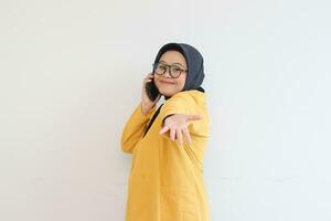 Beautiful young Asian Muslim woman in glasses, hijab and wearing yellow blazer smiling confident and happy with single hands presenting empty space while holding mobile phone photo