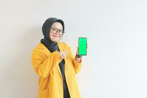 beautiful young asian woman in glasses, hijab and wearing yellow blazer is holding green screen mobile phone with happy face photo