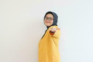 Beautiful young Asian Muslim woman in glasses, hijab and wearing yellow blazer smiling confident and happy with single hands presenting empty space photo